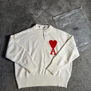 Vit stickad tröja från Ami Paris - Vit stickad tröja från Ami Paris med röd hjärtformad A-logga på bröstet. Tröjan har ribbade muddar vid ärmslut, hals och nederkant samt en klassisk rund halsringning. Perfekt för dig som gillar stilrena detaljer och vill ha något som sticker ut.