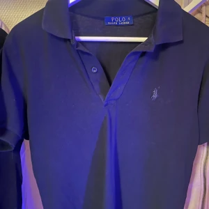 pikétröja från Ralph Lauren - Snygg mörkblå pikétröja från Ralph Lauren med klassisk krage och två knappar framtill. Tröjan har korta ärmar och den ikoniska broderade loggan på bröstet. Perfekt för dig som gillar stilrena och tidlösa plagg.