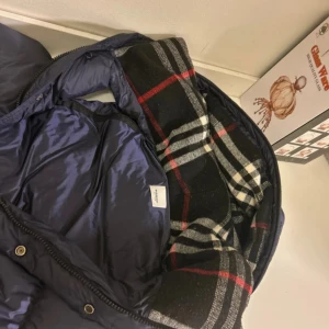 Mörkblå pufferväst från Burberry - Snygg mörkblå pufferväst från Burberry med klassiskt rutigt foder i svart, vitt och rött. Västen har huva, dragkedja och knappar framtill samt två fickor med dragkedja. Perfekt för lager-på-lager och har en riktigt clean look.
