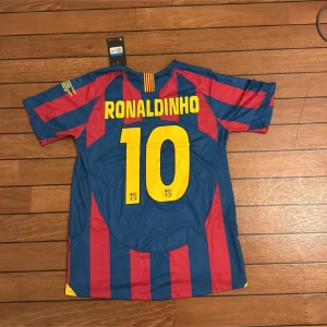 Barcelona Ronaldinho 10 CL Final 2006 tröja - Säljer en officiell FC Barcelona fotbollströja från Champions League-finalen 2006 mot Arsenal, med Ronaldinho och nummer 10 på ryggen. Tröjan är randig i blått och rött, har korta ärmar, broderat klubbmärke och Nike-logga. Materialet är lätt polyester.