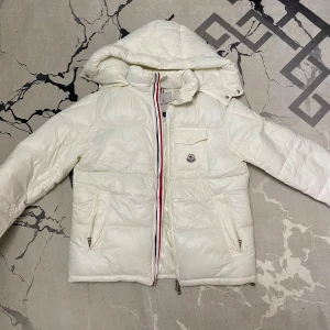 Vit Moncler dunjacka med huva - Säljer en snygg vit dunjacka från Moncler med huva och klassisk logga på bröstfickan. Jackan har dragkedja med rödblå detalj, två sidofickor med dragkedja och en bröstficka med knapp. Materialet är glansigt och fyllt med dun, perfekt för kalla dagar.de går att skanna den och den är helt ny passar båda folk med storlek M Och  L