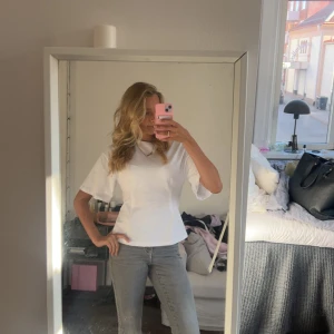 Vit topp med vida ärmar - Säljer en stilren vit topp med rund hals och vida, korta ärmar. Toppen har en lätt figursydd passform och är gjord i ett mjukt bomullstyg. Perfekt att matcha med jeans eller kjol för en clean look.