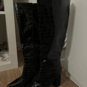 Svarta höga stövlar med klack - Säljer ett par svarta stövlar med krokodilpräglad yta och glansig finish. De har spetsig tå, blockklack och dragkedja på insidan. Snyggt statement till din outfit, passar perfekt till höst och vinter. Materialet är syntet med reptilmönster.