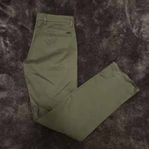 Olivgröna chinos från BOSS 32/32 - Snygga olivgröna chinos från BOSS med regular fit. Byxorna har klassiska fickor, bälteshällor och en diskret metallogo bak. Perfekta för en clean och stilren look. Tillverkade i mjuk bomull för skön känsla hela dagen.