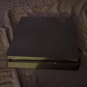 Playstation 4 Slim - Sony Playstation 4 Slim med en vit DualShock 4-kontroll. Konsolen visar mindre kosmetiskt slitage men är i gott skick. Perfekt för gaming och multimedia. Flera USB-portar synliga. Kontroll med mindre repor men fullt fungerande.