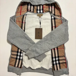 Grå Burberry hoodie med dragkedja - Grå hoodie från Burberry, 10/10 kvalité med klassiskt rutigt foder på insidan av huvan och längs öppningen. Tröjan har hel dragkedja, snörning i huvan och broderad logga på bröstet. Perfekt för de kommande kyliga dagar. Storleken är M, skickas inom 24 timmar. 