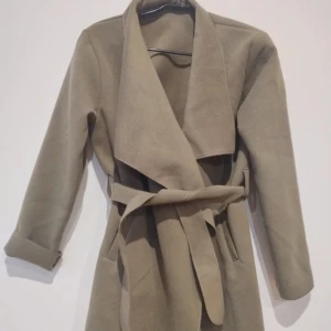 Beige omlott trenchcoat med bälte - Snygg beige trenchcoat med omlott-design och bälte i midjan. Jackan har bred krage och långa ärmar med uppvikta muddar. Perfekt för dig som gillar stilrena och klassiska plagg med modern touch.