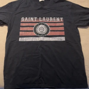 Svart Saint Laurent T-shirt storlek S - Cool svart t-shirt från Saint Laurent med ett stort grafiskt tryck i rött och vitt på bröstet. Trycket har slitna detaljer och texten 'SAINT LAURENT' samt SL-emblem. Klassisk rund hals och korta ärmar. Perfekt för dig som gillar streetstyle och designer. Storlek S