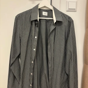 Randig skjorta från Bläck - Snygg svart skjorta med vita vertikala ränder från Bläck. Skjortan har klassisk krage i vitt, knäppning framtill och långa ärmar. Tillverkad i bomull för en skön känsla och stilren look.