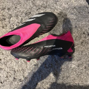 Adidas Predator rosa/svarta fotbollsskor - Säljer ett par Adidas Predator fotbollsskor i svart och rosa med vita detaljer. Skorna har en strumpliknande ovandel utan snörning, mönstrad sula med dobbar och snygga grafiska inslag. Perfekt för dig som vill sticka ut på planen med både stil och funktion.
