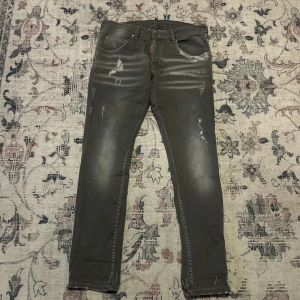 Grå jeans från Dsquared2 med slitningar - Grå jeans från Dsquared2 i storlek 50 med slitna detaljer och diskret tvättad look. Klassisk femficksmodell med DSQ2-logga bak i midjan. Snygg passform och coola slitningar för en trendig vibe. Perfekta för dig som gillar streetstyle och vill sticka ut.