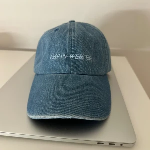 Blå denimkeps från Carin Wester - Säljer en snygg blå keps från Carin Wester i klassisk denim. Kepsen har böjd skärm och vit broderad logga framtill. Perfekt accessoar för en chill och trendig look, passar till både streetwear och casual outfits.