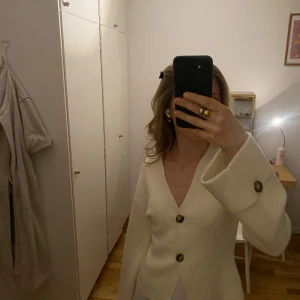 Beige kofta från Gina Tricot med knappar - Superfin beige kofta från Gina Tricot med två stora, mönstrade knappar framtill. Koftan har v-ringning, lång ärm och är gjord i ett mjukt stickat material. Perfekt att slänga över en topp för en chill look.