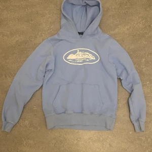 Corteiz Alcatraz Hoodie - Ljusblå hoodie med stor vit tryckt grafik på bröstet, motiv av en byggnad i oval ram. Klassisk huva och magficka, ribbade muddar och nederkant. Mjuk insida, perfekt för chill dagar. Snygg och enkel design som funkar till jeans eller mjukis. Det står S på hoodin fast passar även M och har bara använt den kanske 2 gånger max