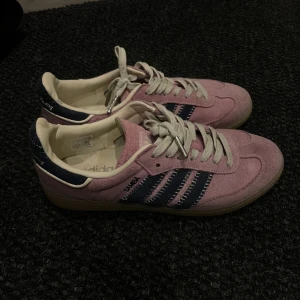 Adidas Samba rosa och marinblå mocka - Säljer ett par Adidas Samba sneakers i rosa mocka med marinblå detaljer och klassiska tre ränder på sidan. Skorna har vit insida, vita snören och gummisula. Perfekt för dig som vill sticka ut med en färgglad och retro stil.