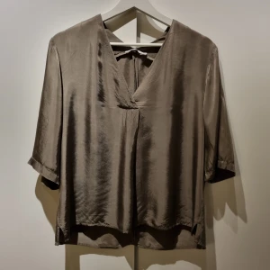 Gråbeige glansig blus från Wera M - Säljer en brun glansig blus från Wera i storlek 38. Blusen har v-ringning, knytning i nacken och trekvartsärmar. Tillverkad i 100% viskos som ger ett lyxigt och mjukt fall. Perfekt för dig som gillar stilrena och enkla plagg med en twist.
