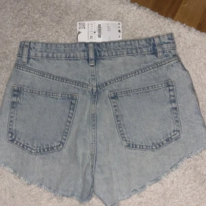 Ljusblå jeansshorts från Zara - Säljer ett par ljusblå jeansshorts från Zara med råa, fransiga kanter och slitna detaljer framtill. Klassisk femficksmodell med hög midja och dragkedja. Perfekta för en chill sommardag.