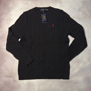 Svart kabelstickad tröja Polo Ralph Lauren - Svart kabelstickad tröja från Polo Ralph Lauren med klassisk rund hals och röd broderad logga på bröstet. Tröjan har ribbade muddar och är långärmad, perfekt för dig som gillar stilren och tidlös design.