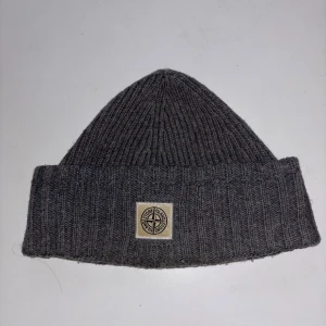 Grå ribbad mössa från Stone Island - Snygg grå ribbstickad mössa från Stone Island med klassisk logopatch framtill. Perfekt för kalla dagar och enkel att matcha med din streetwear eller vinterjacka. Mjuk och skön känsla tack vare ullmaterialet.