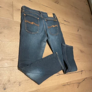 Blå Nudie Jeans 32/32 med orange sömmar - Snygga blå jeans från Nudie Jeans i storlek 32/32. Klassisk femficksmodell med orange kontrastsömmar och Nudie Jeans logga på bakfickan. Jeansen har en rak passform och är tillverkade i slitstarkt denimtyg. Perfekt för dig som gillar stilrena och tidlösa jeans.