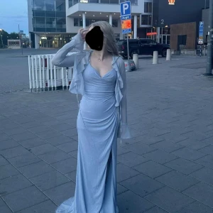Ljusblå glittrig långklänning med snörning - Supersnygg ljusblå långklänning med glittrigt tyg och smala axelband. Klänningen har draperad framsida, snörningsdetaljer i ryggen och en smickrande slits. Perfekt för bal eller festliga tillfällen. Materialet känns mjukt och följsamt. Klänningen har inbyggd bh. Priset går att förhandlas med. Original pris 3400kr i butik.