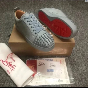 Grå sneakers från Christian Louboutin - Säljer ett par grå sneakers från Christian Louboutin med ikoniska röda sulor och coola nitar på tån. Skorna har snörning och är tillverkade i mocka för en lyxig känsla. Perfekta för dig som vill sticka ut med en edgy och exklusiv stil.