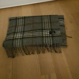 Rutig grå halsduk från A. Christensen - Snygg halsduk från A. Christensen med klassiskt rutigt mönster i grått, vitt och mörkgröna toner. Gjord i merinoull har fransar i ändarna. Perfekt att slänga över axlarna för en chill och stilren look. Kostar 700 ny använd 3 gånger väldigt bra kvalité perfekt för vintern