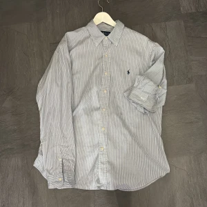 Randig skjorta från Ralph Lauren - Klassisk skjorta från Ralph Lauren i blåvitt randigt mönster. Skjortan har lång ärm, knappar framtill och broderad logga på bröstet. Classic fit-modell i mjuk bomull, perfekt för en clean och stilren look.