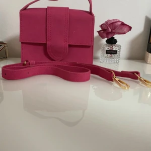 Rosa axelväska i mocka-look - Snygg rosa axelväska med avtagbar och justerbar axelrem i samma färg. Väskan har en stilren, fyrkantig form och stängs med magnetlås. Materialet har en mocka-liknande yta och detaljer i guldmetall. Perfekt för att bära det viktigaste.