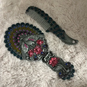 Peacock-inspirerad spegel och kam - Unik handhållen spegel och kam med påfågelsdesign i silverfärgad metall. Båda har detaljerade fjädrar med blå, lila och guld accenter samt rosa blommor och glittriga stenar. Perfekt för dig som gillar detaljer och färg. Spegeln har oval form och kammen är dekorerad med påfågelsmönster.