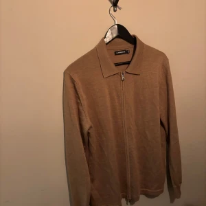 Beige kofta från J.Lindeberg med dragkedja - Snygg beige kofta från J.Lindeberg med hel dragkedja framtill och klassisk krage. Tröjan är stickad i ett mjukt material och har långa ärmar. Perfekt för lager-på-lager och passar till både jeans och chinos. Nypris 1800kr