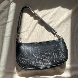 Svart axelväska med krokopräglat mönster - Snygg svart axelväska med krokopräglat mönster och silverfärgad dragkedja. Väskan har en avtagbar axelrem och flera fack inuti för smidig organisering. Perfekt storlek för det viktigaste och ger en cool vibe till din outfit.