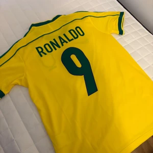 Brasilien Ronaldo 9 fotbollströja Nike M - Gul Brasilien fotbollströja från Nike med gröna detaljer, nummer 9 och Ronaldo på ryggen. Klassisk landslagströja med broderat landslagsmärke och Nike-logga framtill. Tillverkad i polyester, perfekt för dig som älskar fotboll och vill hylla en ikon.