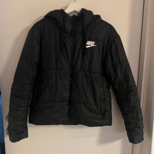 Svart pufferjacka från Nike med huva - Svart pufferjacka från Nike med stor vit logga på ryggen och mindre logga på bröstet. Jackan har huva med snörning, dragkedja framtill och två sidofickor och går att vända ut och in så går att ha på 2 sätt är en vår och höst jacka 