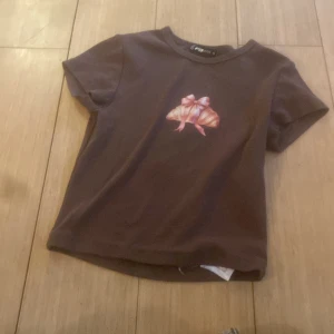 Brun t-shirt med croissanttryck FB Sister - Söt brun t-shirt från FB Sister i storlek S. Framsidan har ett tryck av en croissant med rosa rosett. Klassisk rund halsringning och korta ärmar. Perfekt för dig som gillar unika och lekfulla prints.