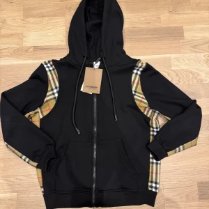 Burberry hoodie - ny med tagg! - Snygg svart hoodie från Burberry med klassiska beige-rutiga paneler längs ärmarna och axlarna. Tröjan har huva med snörning, dragkedja framtill och två stora fickor. Perfekt för dig som vill ha en streetig och lyxig look.