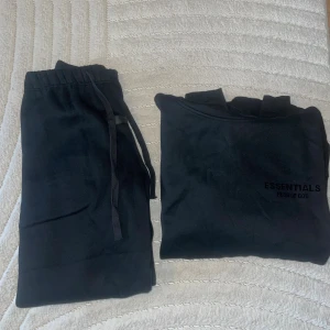 Svart Essentials Fear of God hoodie - Svart hoodie från Essentials Fear of God med diskret logga på bröstet och huva. Tröjan är i mjuk bomull och har en klassisk, avslappnad passform. Perfekt för dig som gillar streetwear och vill ha en stilren look.