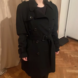 Svart Ullblend trenchcoat med bälte och knappar - Snygg svart ullblend  trenchcoat med klassisk dubbelknäppning, bred krage och bälte i midjan. Jackan har långa ärmar med dekorativa remmar och stora svarta knappar både fram och bak. Perfekt för dig som gillar stilrena och tidlösa plagg. Strl 38 men passar så fint på xs/s se bilderna😊