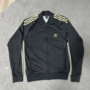 Svart Adidas bomberjacka med guldiga ränder - Snygg svart bomberjacka från Adidas med klassiska guldiga ränder på axlar och ärmar. Jackan har dragkedja framtill, ribbade muddar och Adidas logga i guld på bröstet. Perfekt för dig som gillar sportig och stilren streetwear.