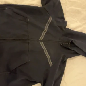 Svart Nike tech från Nike, storlek L - Nike hoodiejacka i svart med hel dragkedja och huva. Jackan har två fickor framtill och diskreta grå ränder över bröstet. Perfekt för chill eller träning, med sportig look och bekväm passform.