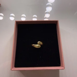 Unik guldpläterad ring med droppform - Säljer en stilren och modern ring i guldpläterad metall med två droppformade detaljer. Ringen har en öppen design som ger en cool och trendig vibe. Perfekt för dig som gillar minimalistiska smycken med en twist.