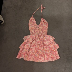 Blommig volangklänning med halterneck - Supersöt kort klänning med blommigt mönster i rosa, rött och gult. Klänningen har halterneck med knyt, omlott-v-ringning och flera volanglager i kjolen. 