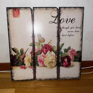 Fin blommig tavla  - . Bilderna visar en dekorativ väggtavla i tre delar med blommotiv och texten 'Love though you have never been hurt before'. Tavlan är i gott skick och har upphängningsanordning på baksidan. Inga synliga defekter eller slitage.