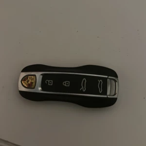 Porsche bilnyckel - Elegant Porsche bilnyckel i svart och silver med tydliga knappar för lås, upplåsning och bagage. Nyckeln har Porsche-emblemet och är i gott skick utan synliga repor eller skador. Perfekt för dig som behöver en extra eller ersättningsnyckel.