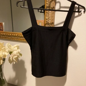 Svart linnetopp med breda axelband - Enkel och stilren svart linnetopp med breda axelband och rak halsringning. Toppen är ärmlös och har en croppad passform som passar perfekt till höga jeans eller kjol. Gjord i mjuk bomull för en bekväm känsla.