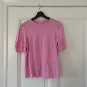 Rosa topp med puffärm - Söt rosa topp med rund halsringning och korta puffärmar. Tillverkad i mjuk bomull, perfekt för vår och sommar. Enkel och stilren design som passar till många olika outfits.