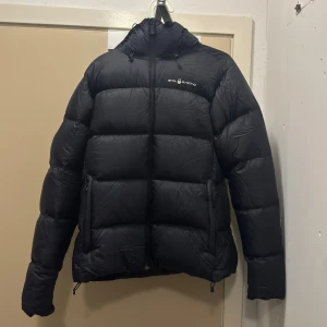 Svart pufferjacka från Sail Racing - Svart pufferjacka från Sail Racing med huva och logga på bröstet samt bak på huvan. Jackan har en stilren look, två sidofickor med dragkedja och en hög krage. Perfekt för kalla dagar och har en riktigt skön passform.