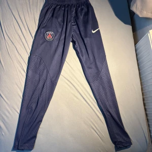 PSG Nike träningsbyxor marinblå - Marinblå träningsbyxor från Nike med PSG-logga på ena benet och vit swoosh på andra. Byxorna har smal passform, elastisk midja och diskret mönster på nedre delen av benen. Perfekta för fotboll eller chill. Materialet är polyester.