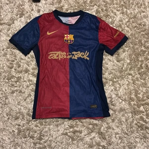 Barça x Cactus Jack Nike fotbollströja - Säljer en Barcelona fotbollströja i samarbete med Cactus Jack och Nike. Tröjan har klassisk halv-röd, halv-marinblå design, guldiga detaljer och klubbmärke på bröstet. Tillverkad i lätt Dri-Fit material som andas, perfekt för match eller träning.