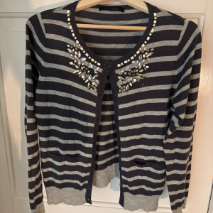 Randig kofta med pärlor från Cynical - Snygg kofta från Cynical i mörkblått och grått med horisontella ränder. Rund halsringning dekorerad med pärlor och glittriga stenar vid axlarna. Långärmad modell med knappar framtill och små fickor. Perfekt för dig som gillar detaljer och mönster.